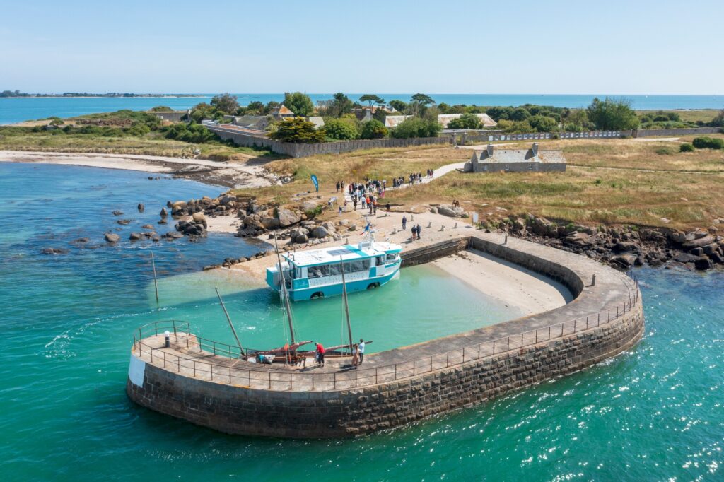 Le bateau amphibie est dans le port de Tatihou. La mer est turquoise et on voit les visiteurs sur le chemin qui mène à l'intra-muros